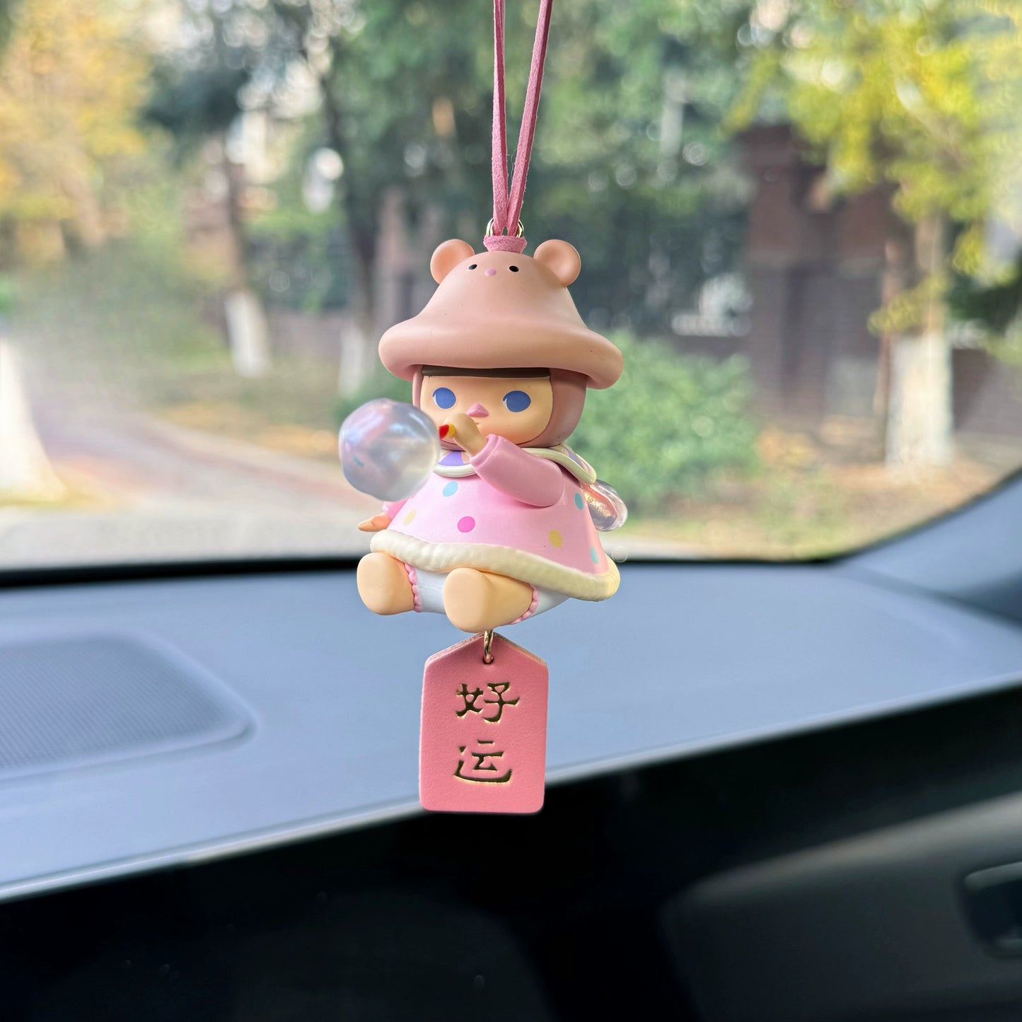Luvbubu BubbleBear charm doll in pink outfit with bear hoodie and crystal ball, hanging on car dashboard with Lucky tag